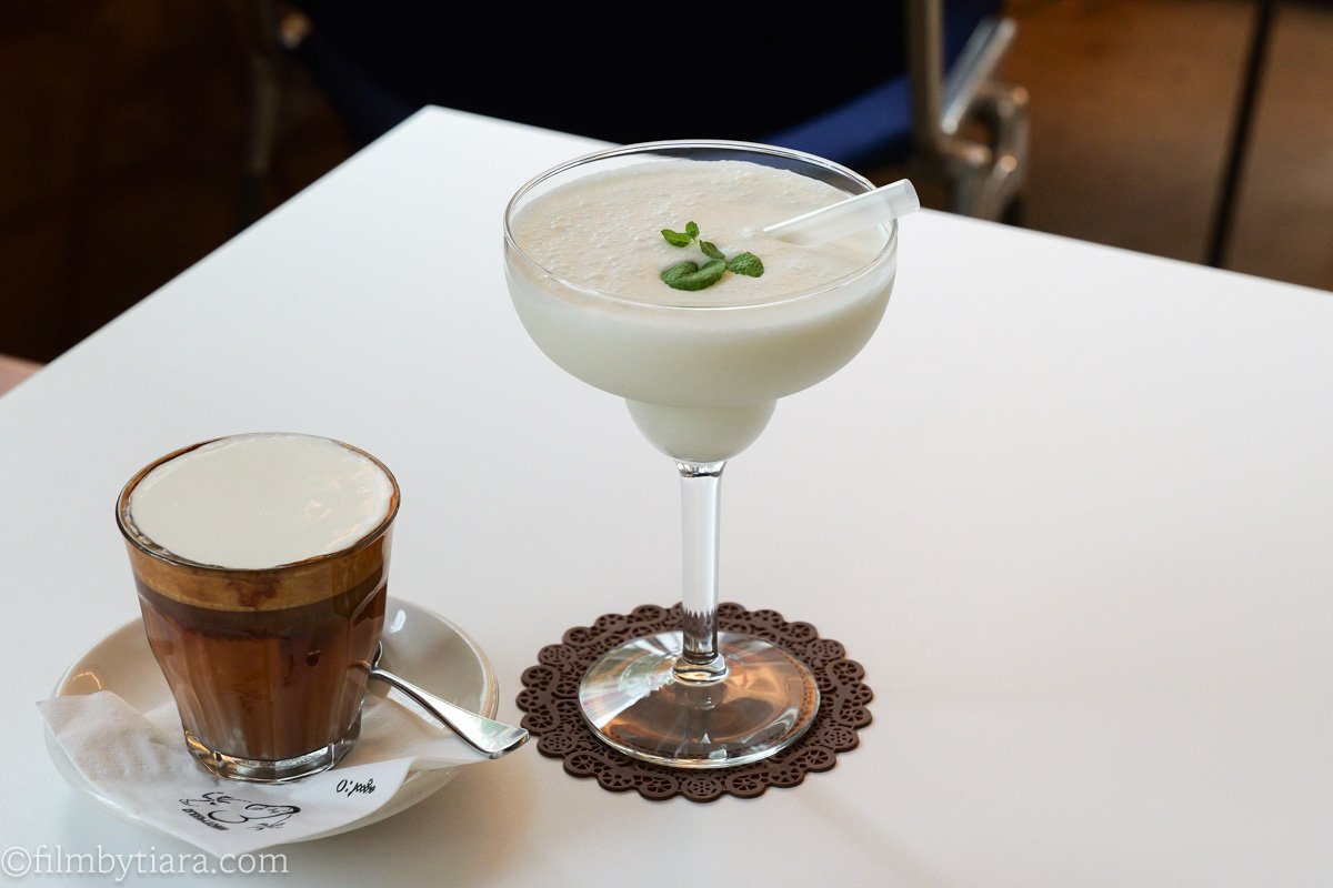 A cream-topped coffee drink next to a non-alcoholic piña colada garnished with mint on a white cafe table