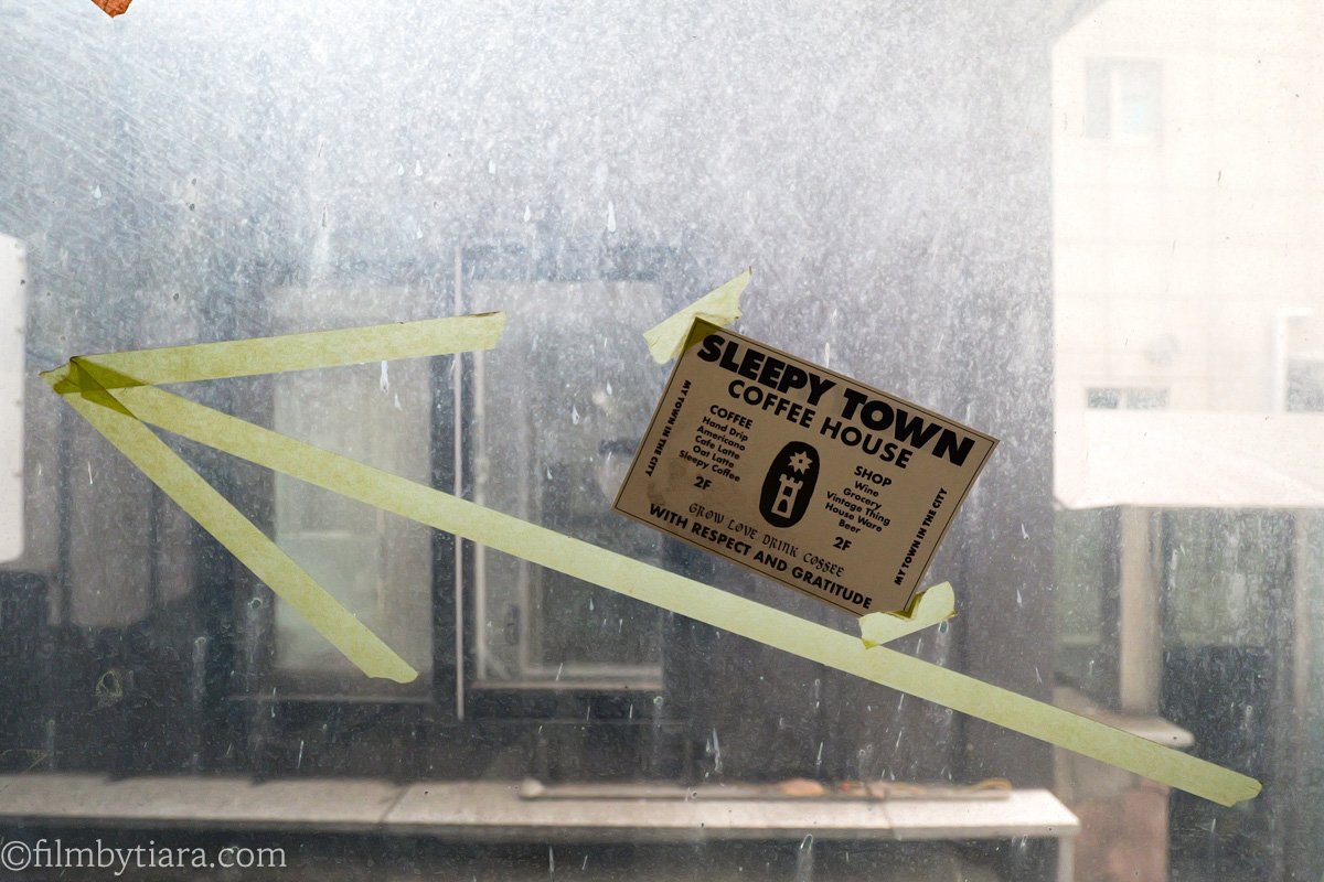 Taped paper sign listing Sleepy Town's coffee and grocery menu on a rain-streaked window