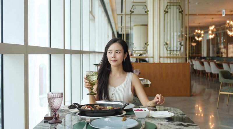 Woman seated at a window table at Restaurant Voisin holding a glass, with the ribeye course on the table